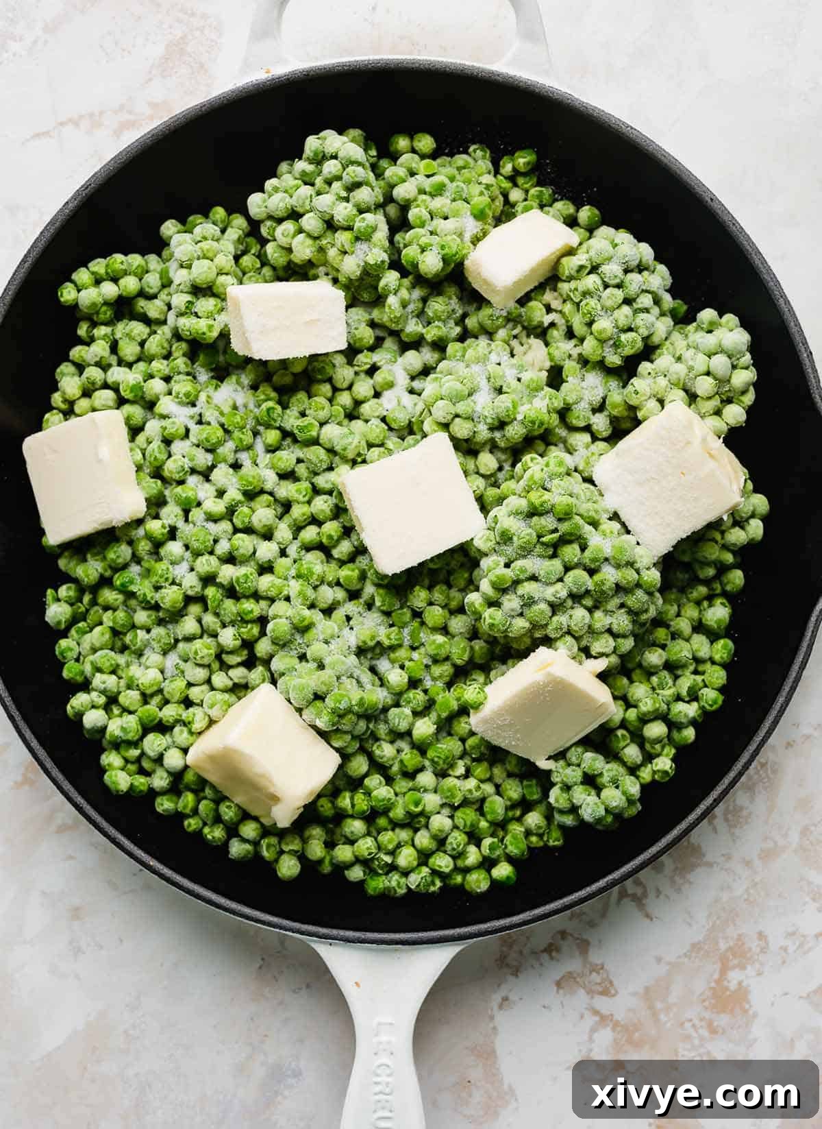 Elevate Your Frozen Peas 4 A cast iron skillet filled with frozen peas, featuring cubes of unsalted butter, granulated sugar, and minced garlic scattered on top, ready for cooking.