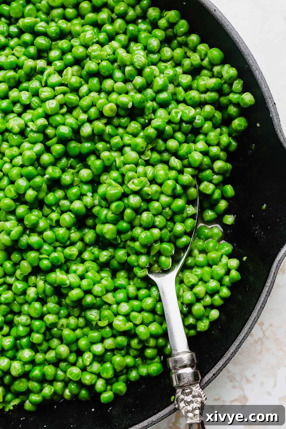 Elevate Your Frozen Peas 6 A close-up shot of perfectly cooked green peas being scooped from a skillet with a silver serving spoon, ready to be enjoyed as a delicious side dish.