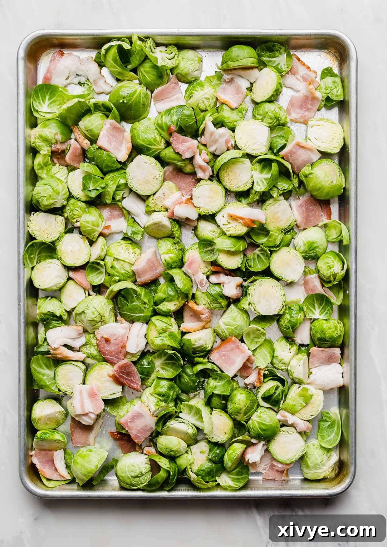 Unbaked Brussels sprouts and raw bacon chunks on a baking sheet.