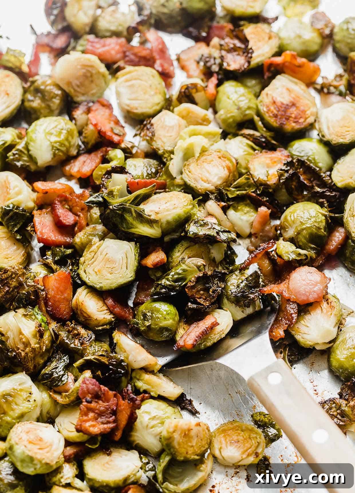 A metal spatula scooping off Roasted Brussels Sprouts with Bacon from a baking sheet.