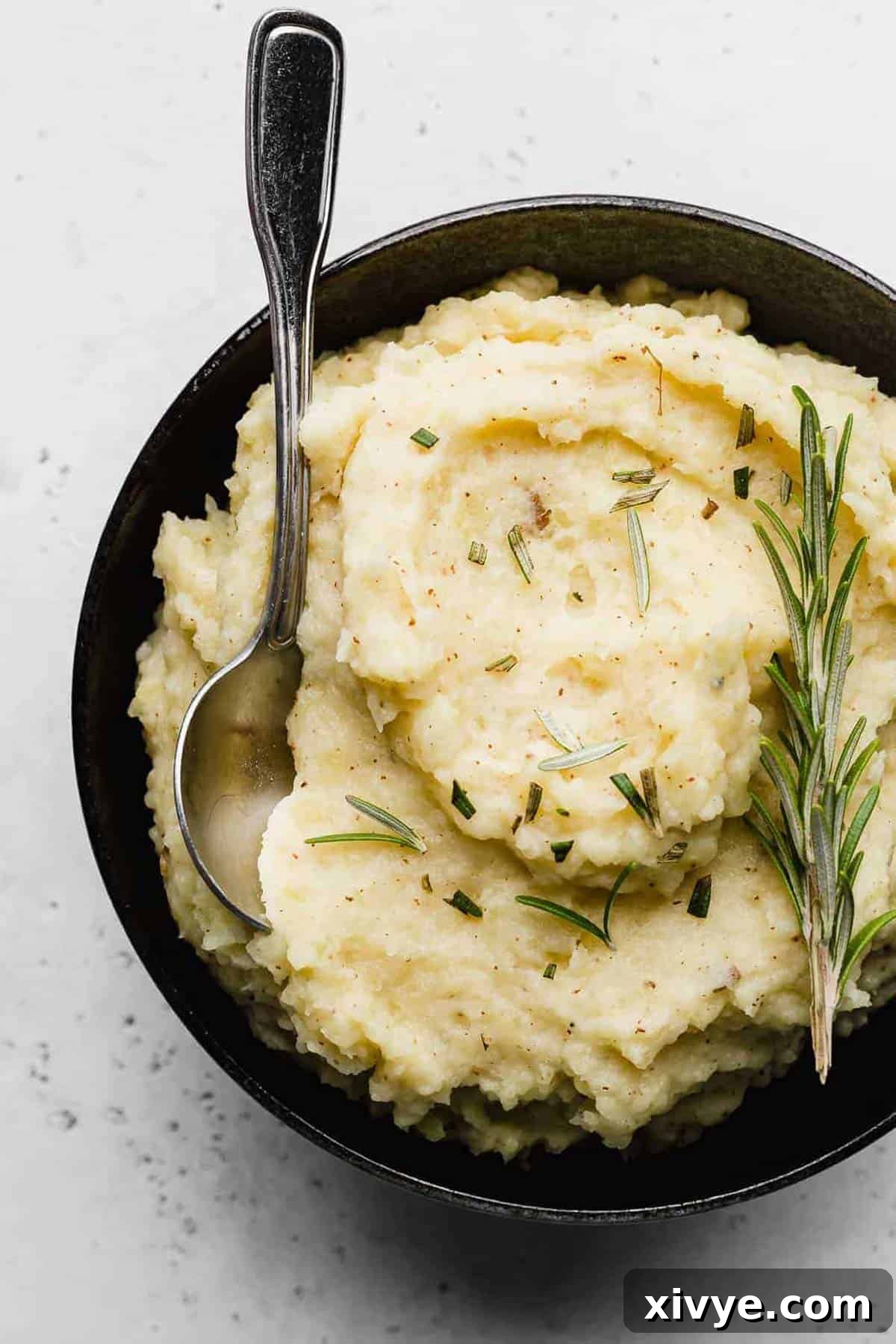 A spoon nestled in a black bowl that's filled with creamy Rosemary Brown Butter Mashed Potatoes, garnished with a sprig of rosemary.