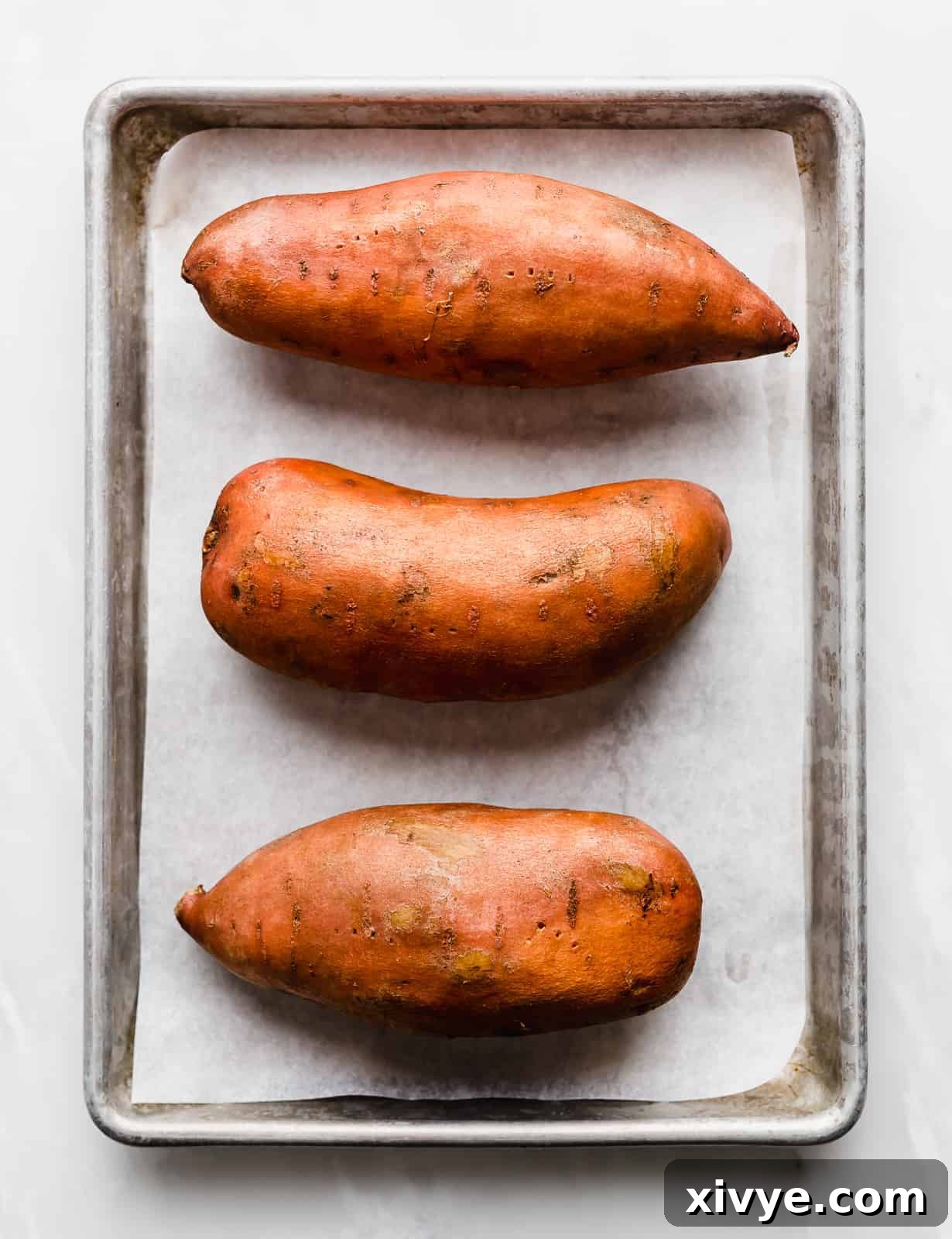 Golden Baked Sweetness 4 Three large sweet potatoes on a white parchment lined baking sheet.