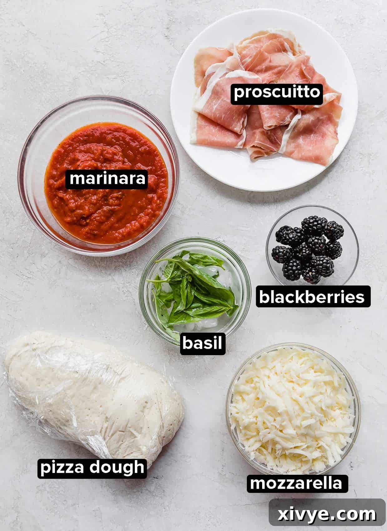 Blackberry Ricotta Pizza ingredients laid out on a gray background, including pizza dough, cheese, basil, fresh blackberries, prosciutto, and a bowl of marinara sauce.