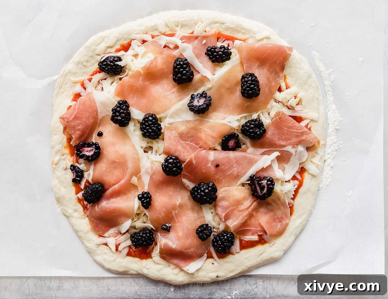 An unbaked pizza topped with prosciutto, fresh blackberries, and mozzarella cheese, ready for the oven.