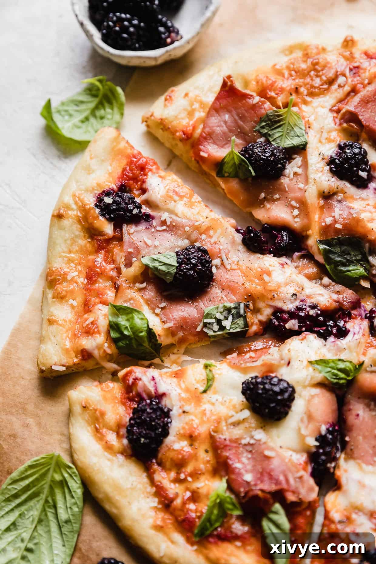 A close-up of a slice of Blackberry Pizza, showcasing the melted cheese, prosciutto, and vibrant blackberries, garnished with basil.