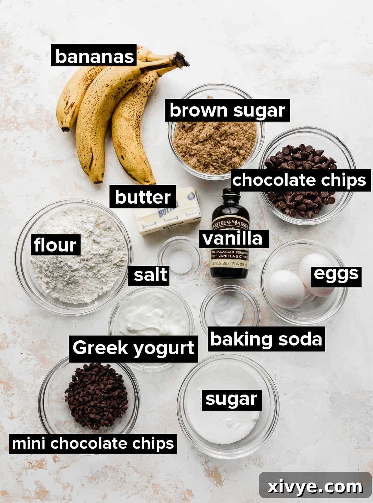 Collection of essential ingredients for chocolate chip banana bread, including ripe bananas, eggs, flour, sugars, yogurt, and chocolate chips, neatly arranged on a pristine white surface.
