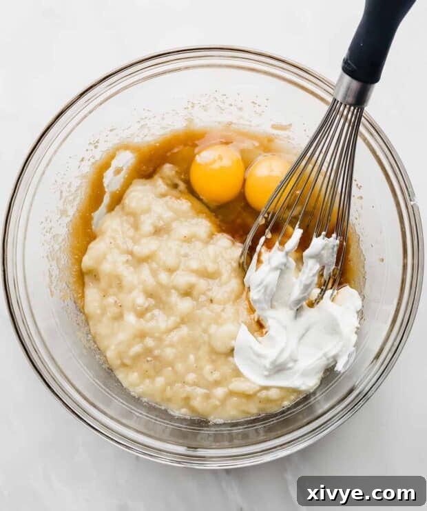 Mashed bananas, Greek yogurt, and eggs added to the sugar and butter mixture in a glass bowl.