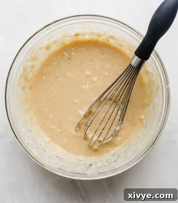 A whisk stirring a beautifully combined banana bread batter in a glass bowl, showing a smooth and creamy consistency.