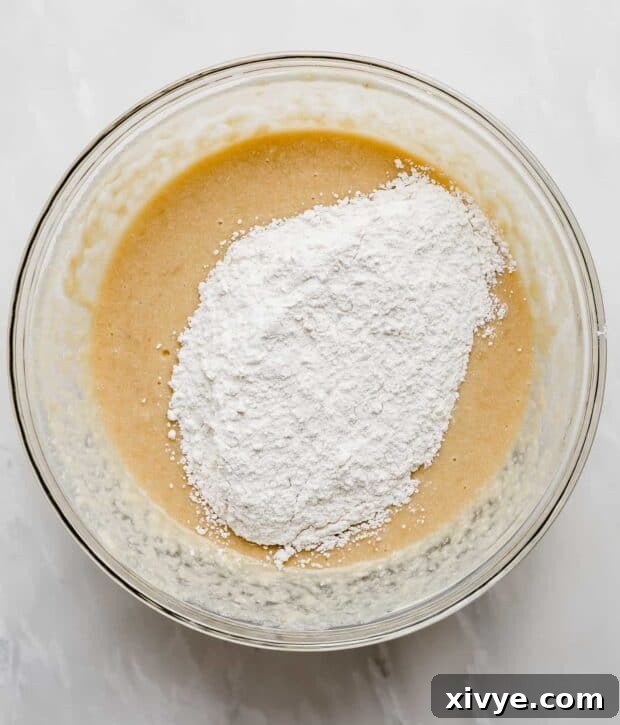 Flour being carefully poured over the banana bread batter in a glass bowl, ready for gentle mixing.