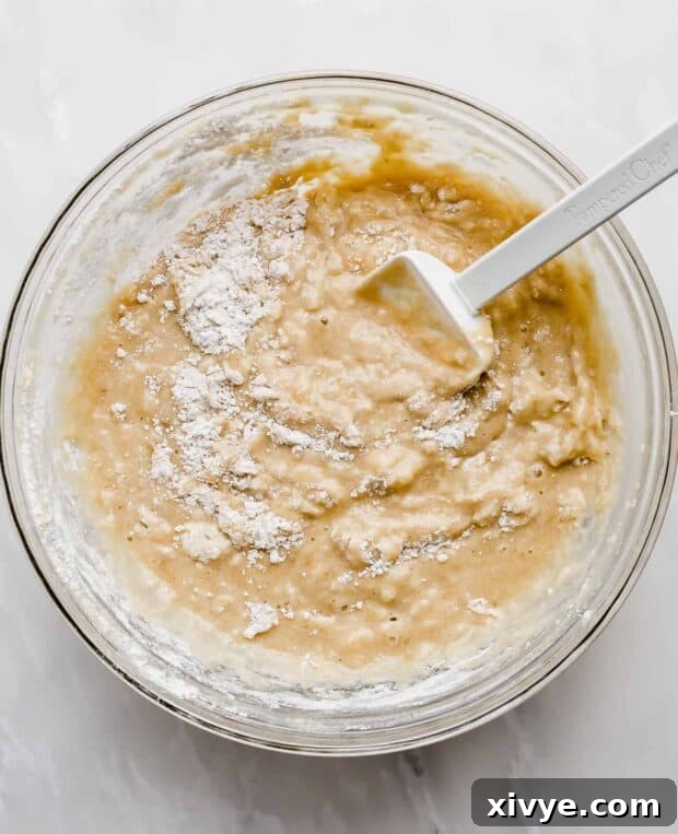 A spatula gently folding dry ingredients into banana bread batter in a glass bowl, showing the consistency when just combined.