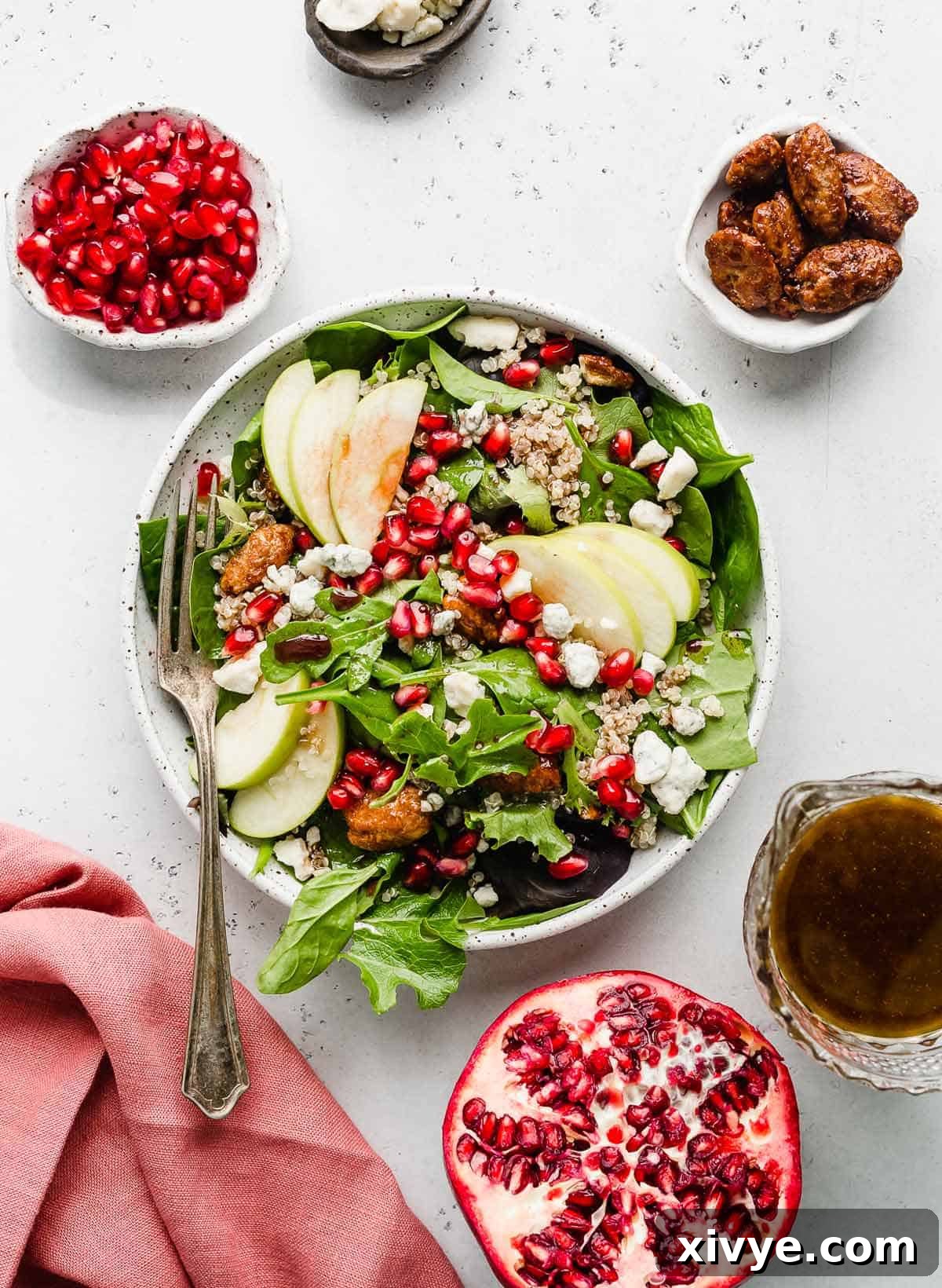 A festive Pomegranate Salad artfully arranged on a white background, surrounded by whole pomegranates, shelled nuts, a red napkin, and a glass of pomegranate dressing.