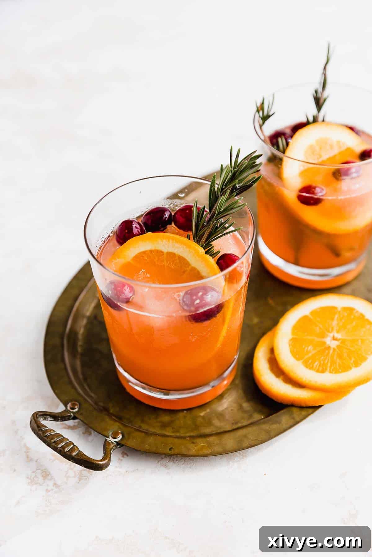 A glass filled with sparkling Christmas Punch, garnished with fresh cranberries and orange slices, sits on a rustic bronze oval tray, surrounded by festive greenery and twinkling lights, capturing the cozy essence of holiday entertaining.