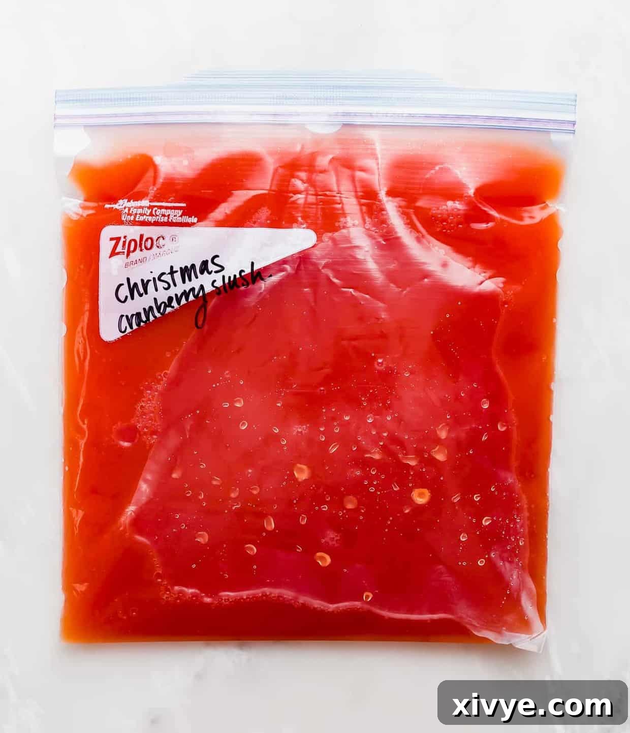 A clear gallon-size freezer bag, filled with a vibrant red juice mixture, lies flat in a freezer, showcasing the essential first step of preparing the cranberry and orange juice blend for making Christmas Slush.