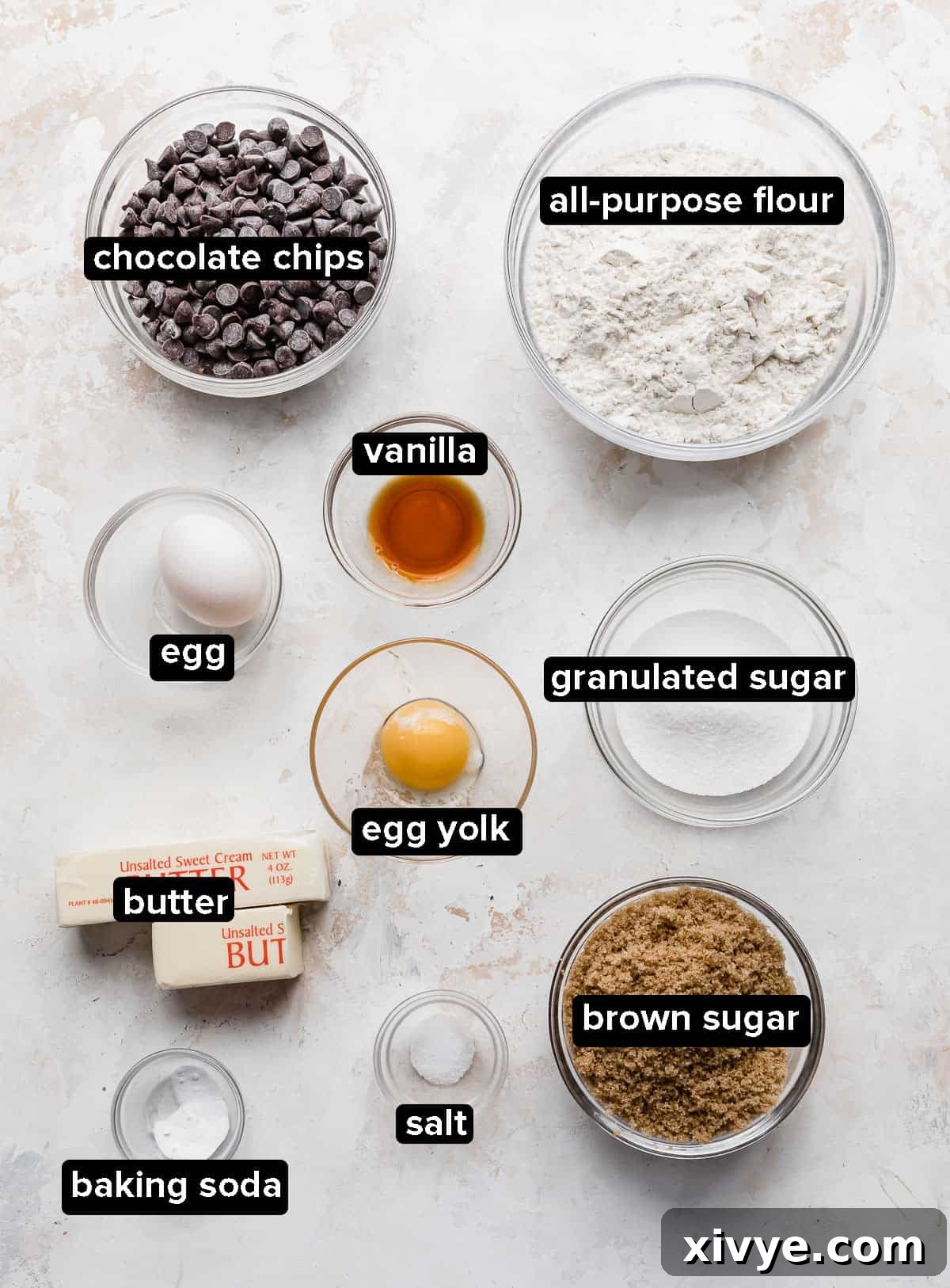 Chocolate Chip Cookie Bar ingredients in small glass bowls on a white textured background.