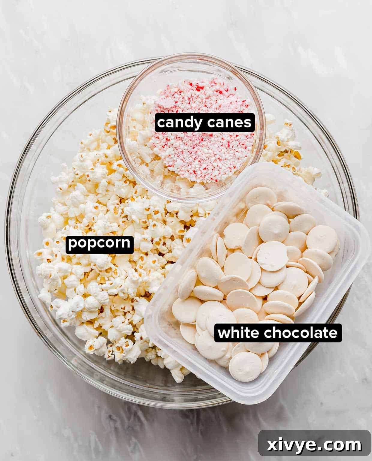 Close-up shot of the simple ingredients for Candy Cane Popcorn laid out on a clean white background: fluffy popcorn, glistening crushed candy canes, and pristine white chocolate wafers.