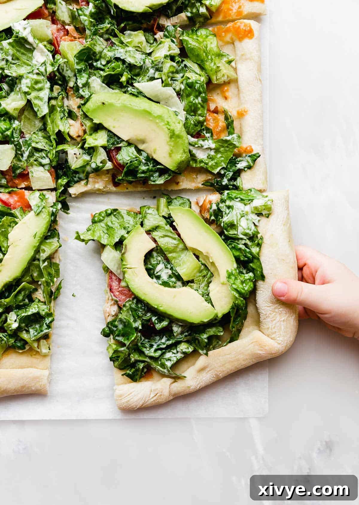 A hand grabbing a square slice of CPK Club Pizza that's topped with fresh avocado slices and lettuce.