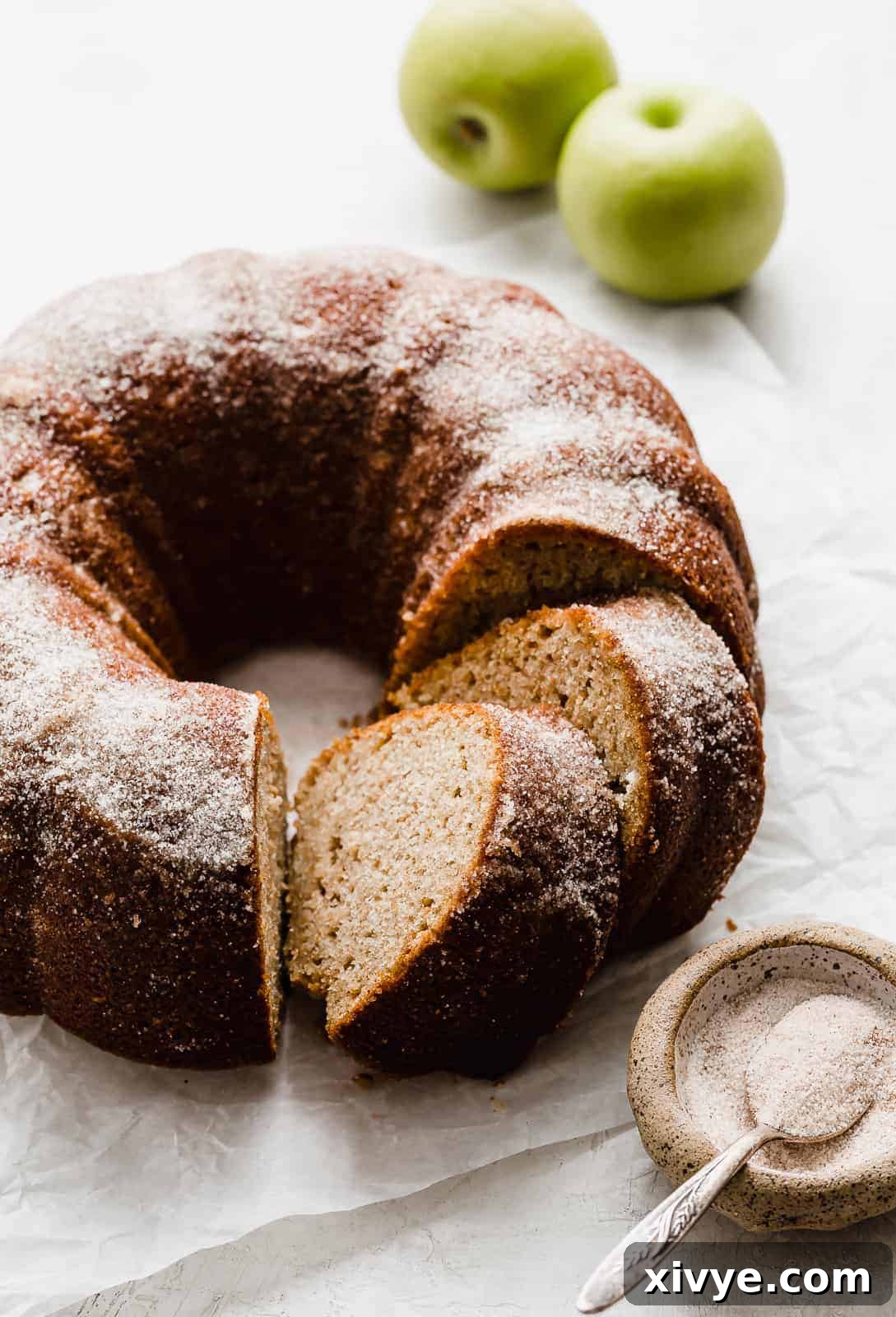 An exquisitely baked Apple Cider Bundt Cake, generously dusted with sparkling cinnamon sugar, presented invitingly on a white cake stand.