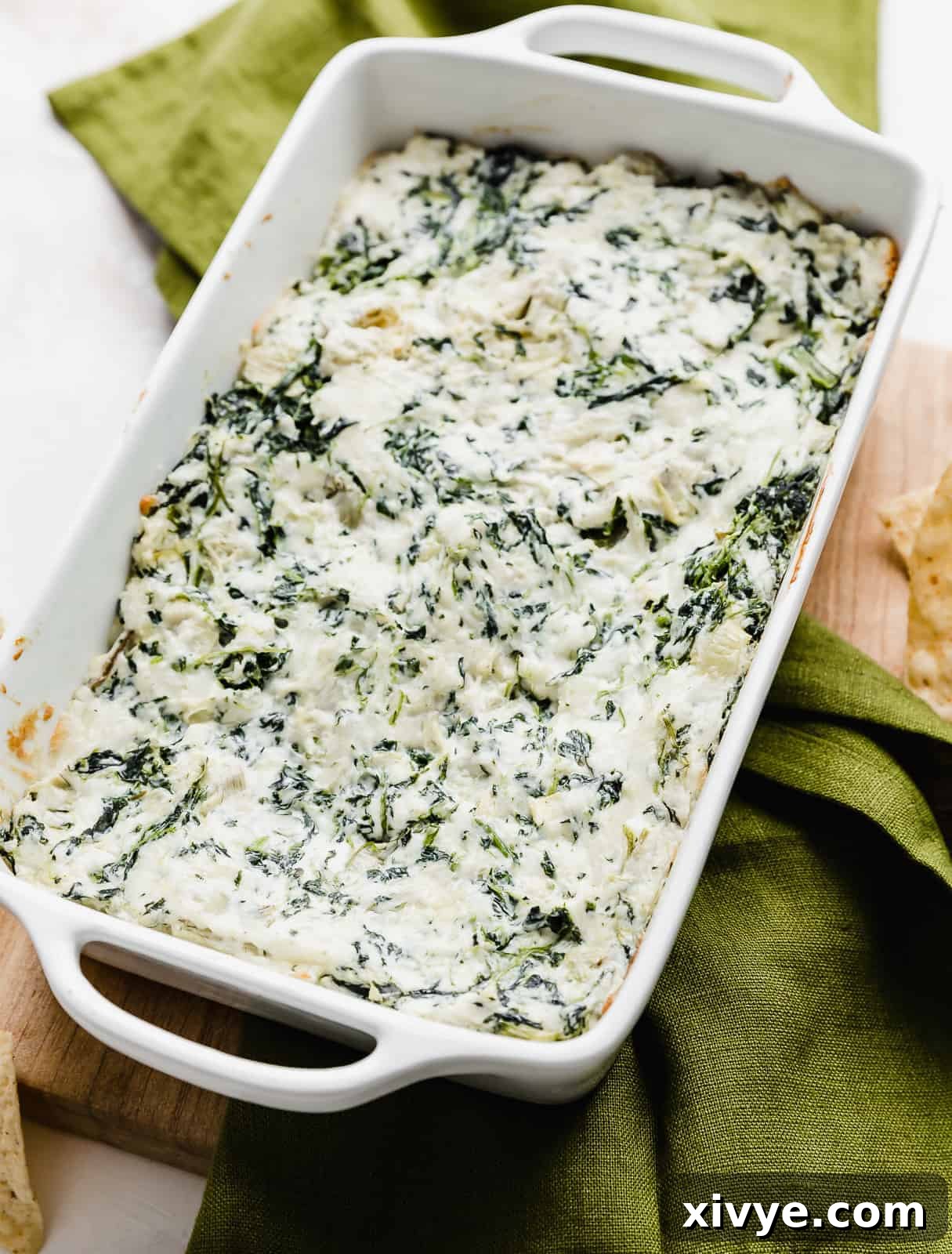 Sizzling Spinach Artichoke Delight 6 A freshly baked hot spinach artichoke dip in a white baking dish, elegantly placed on a dark green napkin, ready for serving at a gathering.