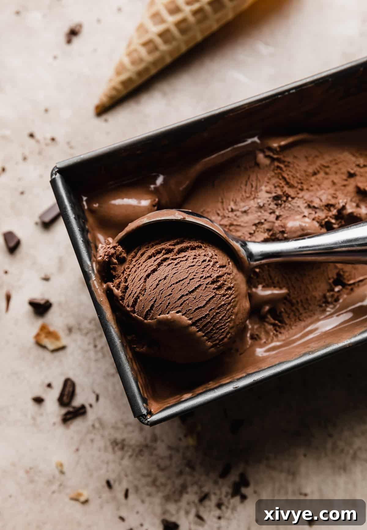 An ice cream scoop rolling out a portion of Homemade Dark Chocolate Ice Cream from a square bread pan, showcasing its rich, dark color and irresistibly creamy texture.