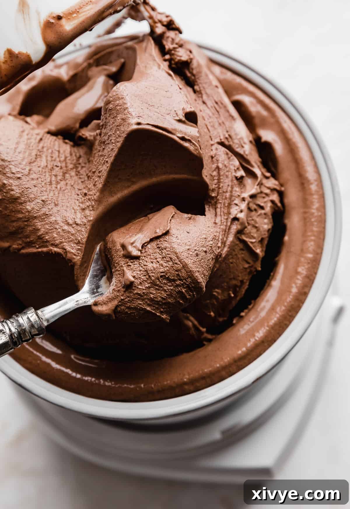 Creamy dark chocolate ice cream being pulled from an at-home ice cream maker, with a spoon scooping out some of the ice cream, showcasing its soft-serve consistency and rich color.