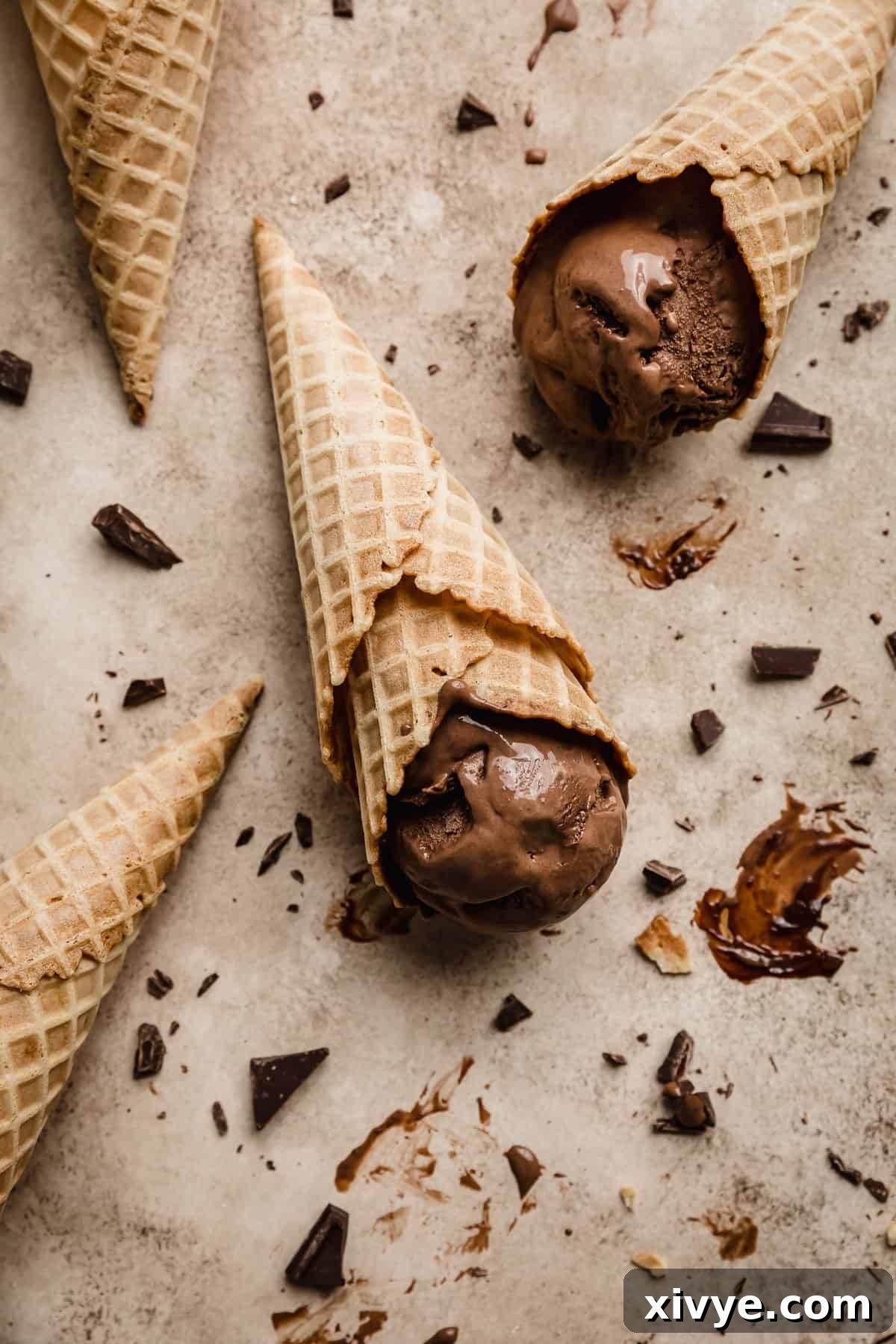 Homemade Dark Chocolate Ice Cream in a waffle cone laying flat on a light brown textured background with chopped chocolate scattered around, signifying a delightful treat ready to be enjoyed.