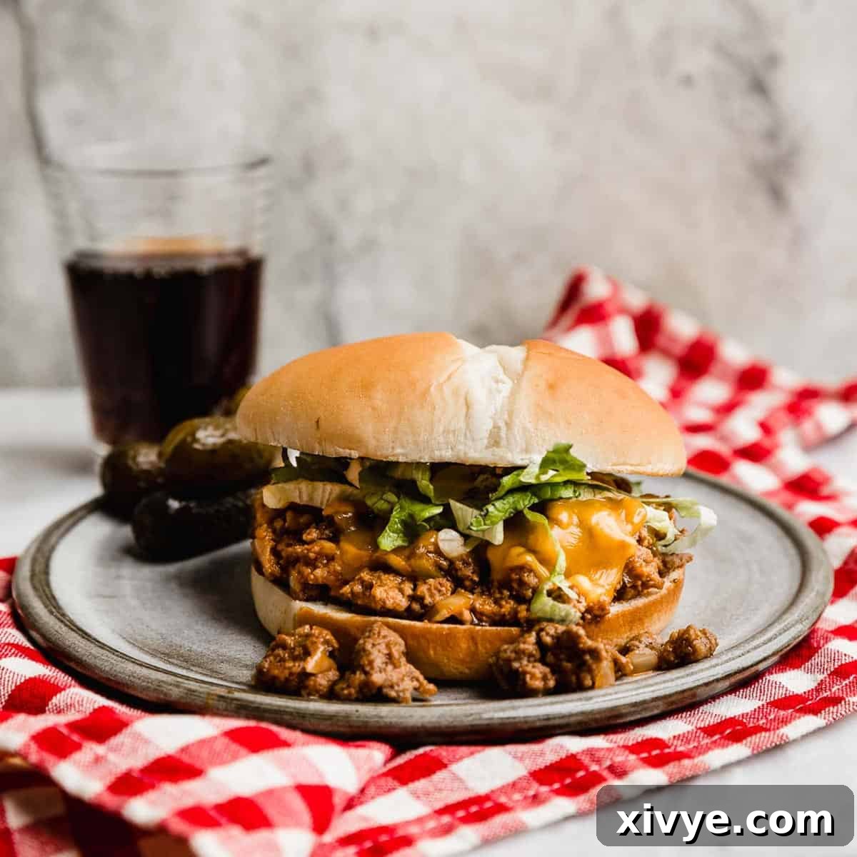 A Cheeseburger Sloppy Joes recipe on a bun with cheese and shredded lettuce with a glass of brown soda in the background.