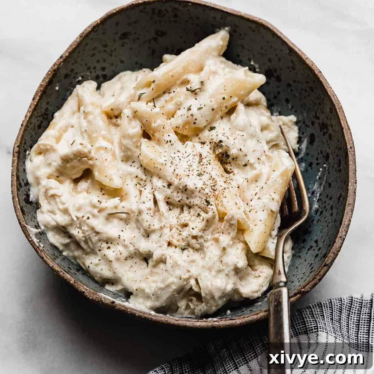 A bowl of creamy chicken Alfredo made tin penne noodles in a bowl on a white marble surface.