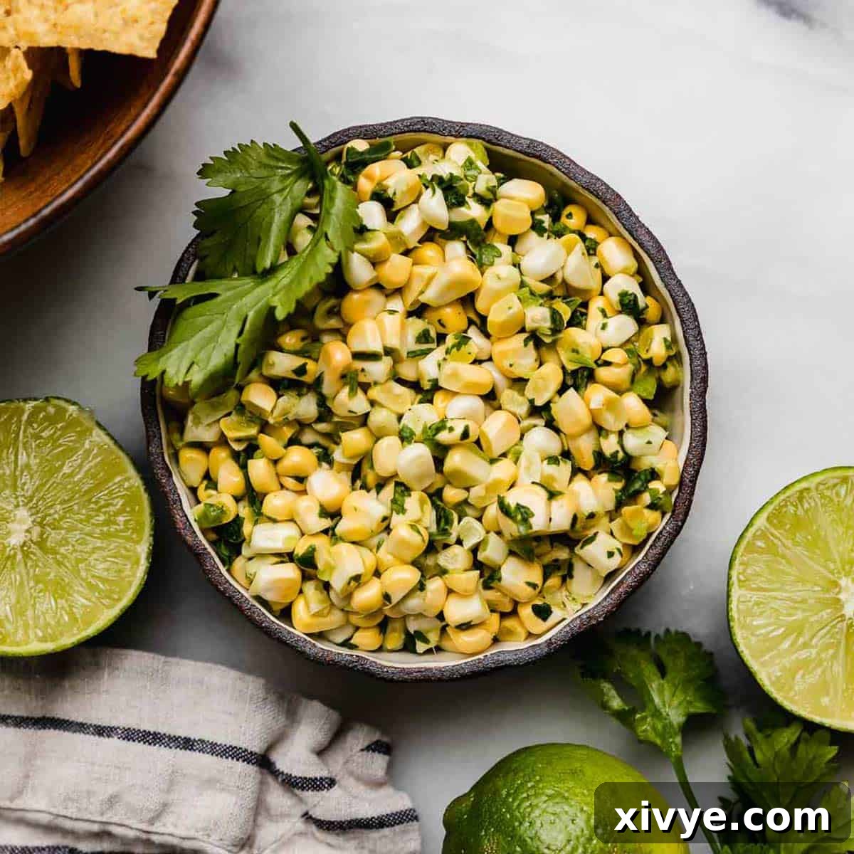 Overhead photo of corn salsa made with raw corn in a black bowl on a white background with fresh limes surrounding the bowl.