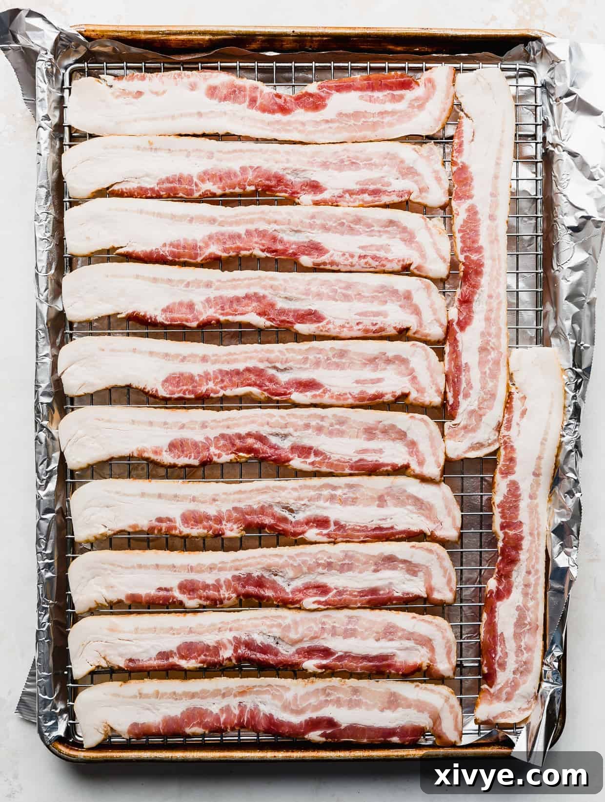 The Secret to Perfectly Crispy Oven Bacon 4 Raw bacon strips neatly arranged in a single layer on a wire rack, which rests inside a foil-lined baking sheet, ready for the oven.