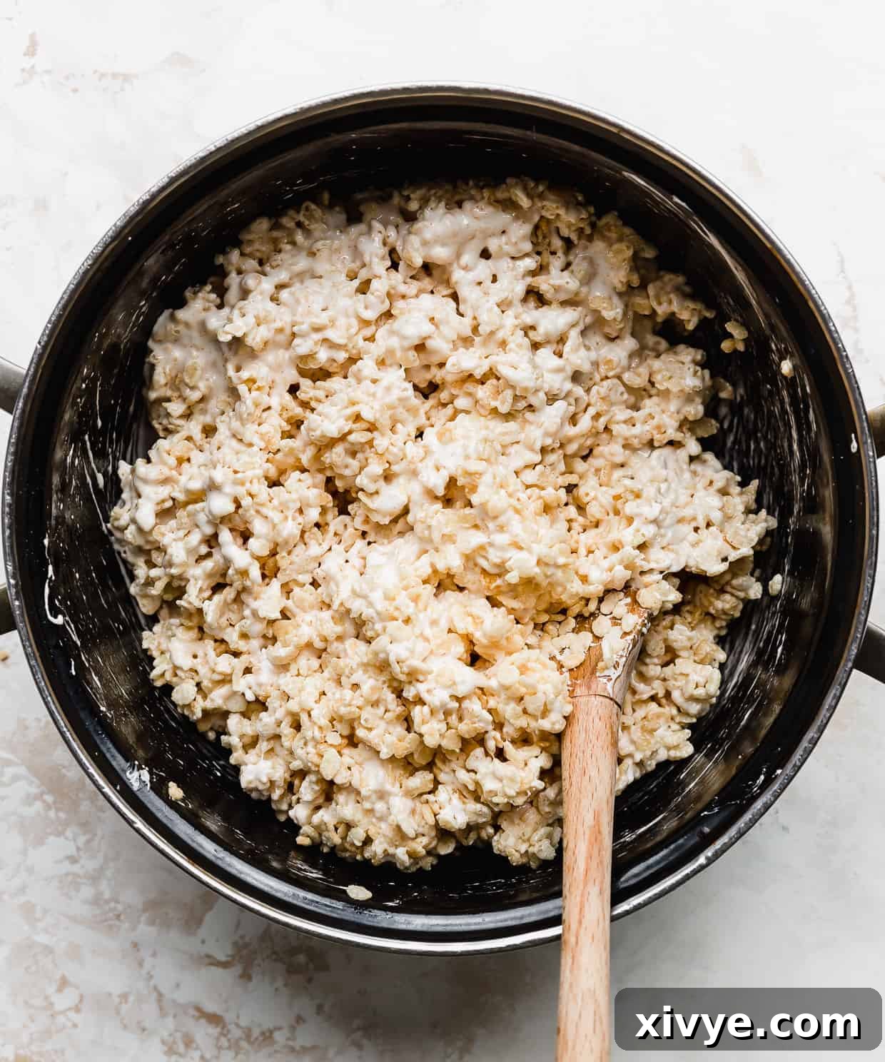 A large black pot against a white background with a wooden spoon stirring Rice Krispie cereal into melted marshmallows.