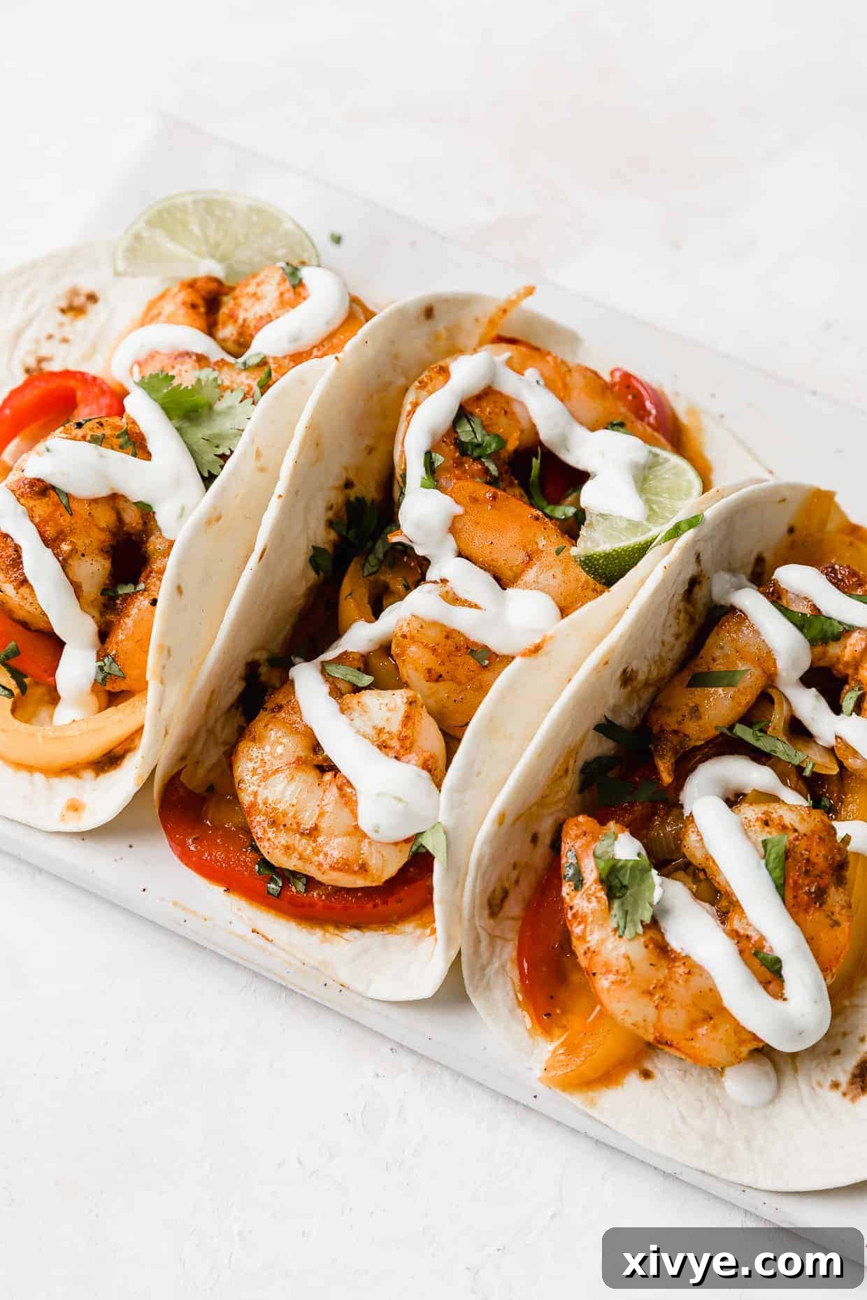Sheet Pan Fiesta Shrimp 6 Three beautifully assembled shrimp fajitas, artfully drizzled with a smooth white lime crema, presented on a minimalist white plate for an appetizing display.